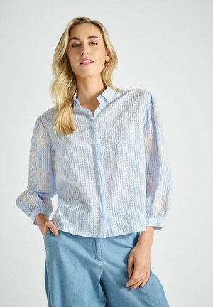 Blonde vrouw draagt een lichtblauwe blouse met een getextureerd patroon en doorschijnende bloemenmouwen, gecombineerd met loszittende blauwe broek, poseert met één hand in de zak.