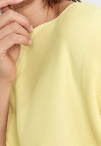 Gul stickad topp med mjuk textur, med en vid halsringning och ribbad detaljering. Nærbild visar en hand som försiktigt rör vid tyget.