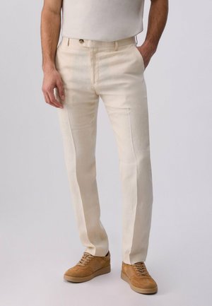 Hombre con pantalones casuales beige y zapatillas de tono marrón claro, de pie con una mano en el bolsillo contra un fondo liso.