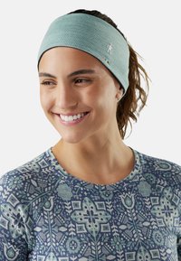 Jeune femme souriante portant un haut bleu-vert à motifs et un large bandeau bleu clair, les cheveux attachés en queue de cheval.
