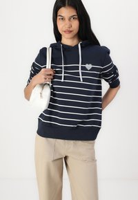 Niebieska bluzka z kapturem w paski, z białymi paskami, z małym detalem w kształcie serca. Połączona z beżowymi spodniami i białą torbą na ramię. Teksturowany materiał.