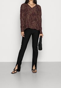 Bordeaux farbener langärmliger V-Ausschnitt-Pullover mit einem wellenförmigen Muster, kombiniert mit schwarzen hochgekrempelten Hosen mit seitlichen Schlitzen und schwarzen Sandalen mit Riemchen.