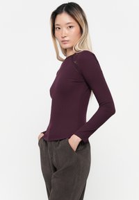 Langsleeve, paarse ribgebreide top met een kantaccent op de schouder, gecombineerd met bruin corduroypantalon. Glad textuur en getailleerd ontwerp.