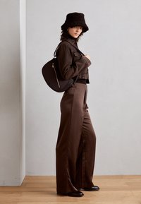 Braune Jeansjacke im cropped Schnitt, braune weit geschnittene Hose, schwarzer flauschiger Bucket Hat und eine schwarze Handtasche mit geschwungenem Design und goldenen Beschlägen. Glatte Texturen.