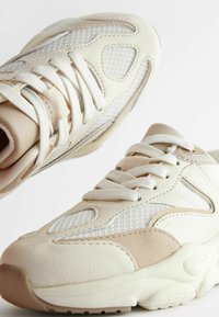 Baskets beiges et blanc cassé avec des panneaux en maille, des accents en cuir texturé, bout rond et lacets plats. Semelle en caoutchouc épaisse pour un soutien.