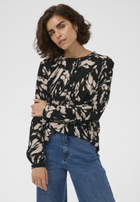 Haut à manches longues avec un motif noir et beige, fermeture à boutons sur le devant, tissu texturé et coupe décontractée, porté avec un jean bleu.