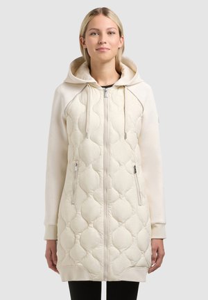 Jonge vrouw die een lange, crème-kleurige gewatteerde jas met capuchon draagt, voorzien van een ritssluiting aan de voorkant en ritszakken, staand tegen een effen achtergrond.