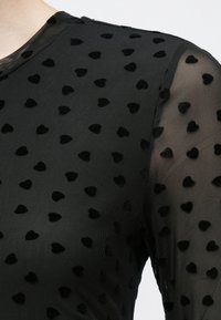 Melnas acs tops ar teksturētām sirsnveida polka punktiem, gluda virsma un pieguļošs dizains. Plānas, garas piedurknes.