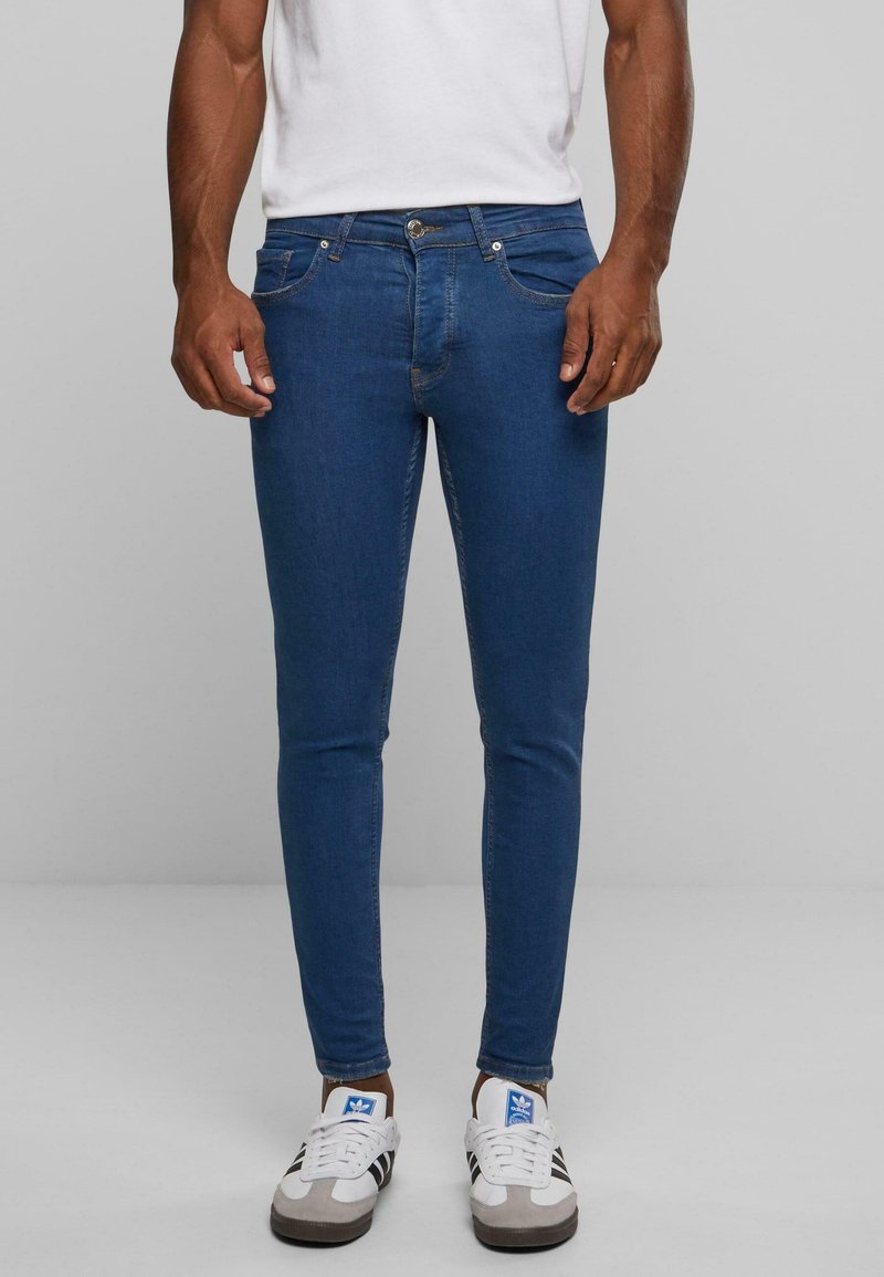 Jeans de mezclilla ajustados en azul oscuro, con cinco bolsillos, un cierre de botón y costuras en contraste. Combinados con zapatillas blancas y grises.