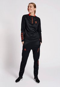 Czarna sportowa koszulka z długim rękawem z pomarańczowymi akcentami i zamkiem błyskawicznym, w zestawie z czarnymi spodenkami dresowymi z pomarańczowymi detalami i logo.