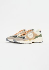 Beige, zöld és szürke velúr keverékéből készült sneakers, narancssárga fűzőkkel, masszív fehér talppal, és feltűnő logóval az oldalon.