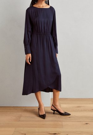 Frau in einem marineblauen, langärmligen Midikleid mit gerafftem Bund und schwarzen, spitzen Slingback-Absätzen, die auf einem Holzboden steht.