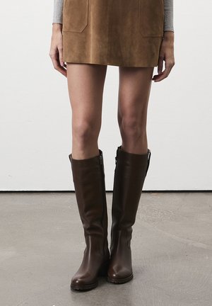 Bottines marron à hauteur de genou en cuir lisse, avec un détail de fermeture éclair sur le côté et une semelle en caoutchouc texturée. Portées avec une jupe en daim marron.