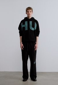 Σετ μαύρης φούτερ μπλούζας με κουκούλα και παντελόνι φόρμας με μεγάλα πράσινα γράμματα "HU" και στα δύο κομμάτια. Το ύφασμα φαίνεται μαλακό· μινιμαλιστικός σχεδιασμός με άνετη εφαρμογή.