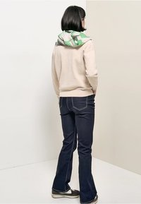Beige hoodie met groene patroonkap, gecombineerd met donkerblauwe uitlopende jeans. Model staat met de rug naar voren, draagt donkere schoenen.