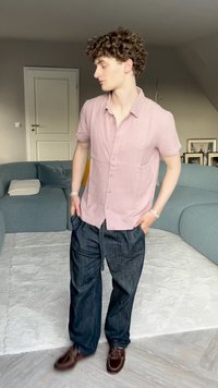 Lys pink kortærmet skjorte med knapper, mørkeblå løstsiddende jeans og brune sko, stående på et lysfarvet tæppe i et moderne interiør.