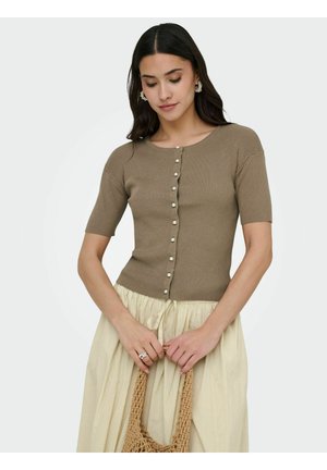 Kvinde med mørkt hår iført en brun ribbet kortærmet knapbluse og et lysebeige skørt, der holder en flettet taske foran en ensfarvet baggrund.