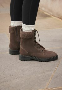Botines de ante marrones con un cuello de piel texturizada, parte delantera con cordones y suela de goma negra, combinados con calcetines claros de canalé y leggings negros.