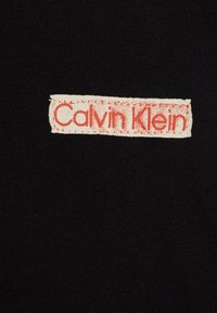 Una camiseta de tela negra presenta un logotipo rectangular texturizado en crema con letras rojas desgastadas que dicen "Calvin Klein" en el pecho.