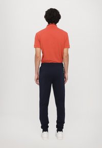 Oranžs polo krekls ar apkakli, apvienots ar tumši ziliem sporta šortiem. Biksēm ir regulējama josta un ribotās malās, tās valkā ar baltām kedām.