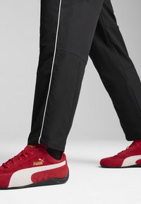 Røde ruskinds-sneakers med hvide detaljer og sorte såler, parret med sorte bukser med hvide sidestriber og en struktureret finish.