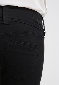 Gros plan sur la poche arrière d'un jean en denim noir avec une double couture et une petite étiquette de marque près de la ceinture, porté avec une chemise blanche.