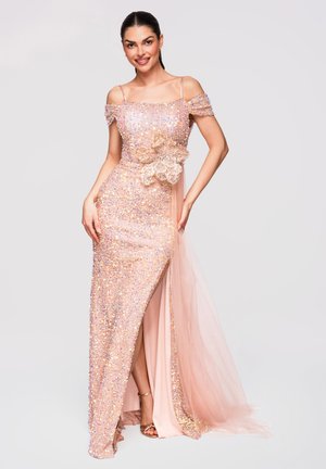 Lachende vrouw in een sprankelende roze off-shoulder jurk met een grote bloemapplicatie en een hoge split tot aan de dij, poserend tegen een effen achtergrond.