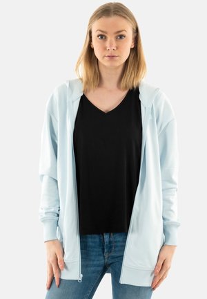 Felpa azzurra con zip e cappuccio, polsini a costine e tasche, indossata sopra una maglietta nera a collo a V, abbinata a jeans blu.