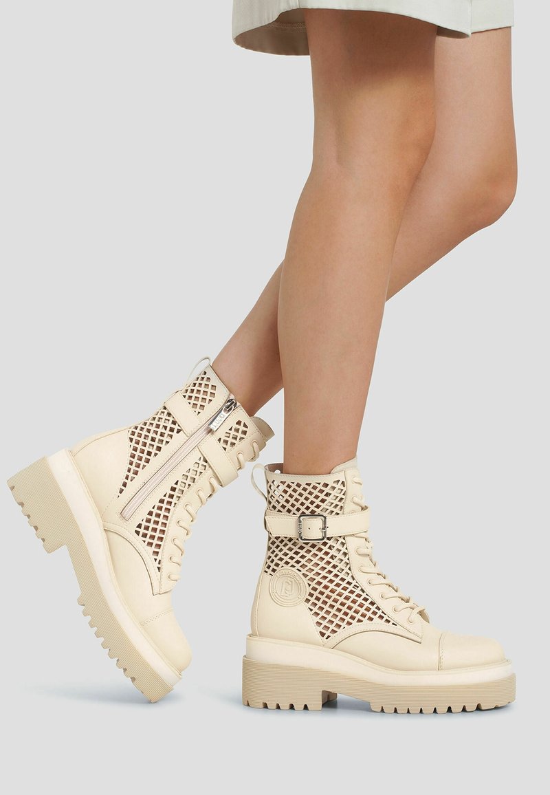 Bottines beiges à lacets avec semelles épaisses et côtés en mesh, portées par une personne debout avec une jambe croisée sur l'autre.