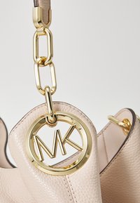 Hellbeige Lederhandtasche mit einem goldenen MK-Logo-Anhänger, ausgestattet mit einer strukturierten Oberfläche und goldenen Hardware-Akzenten.