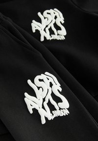 Čierna tkanina s hladkou textúrou má výrazné biele logo. Text loga je vlnitý a znie "ALSA NTS LONDON" na spodnej strane.