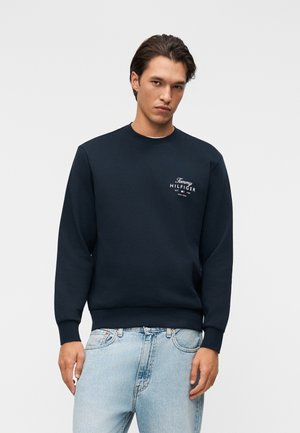 Ung mand iført en mørkeblå Tommy Hilfiger sweatshirt og lyseblå jeans, stående mod en ensfarvet lys baggrund.