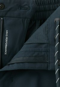 Gros plan sur un pantalon en tissu foncé avec fermeture éclair, bouton, ceinture élastique et l'inscription "MNG PERFORMANCE" imprimée sur la doublure intérieure.
