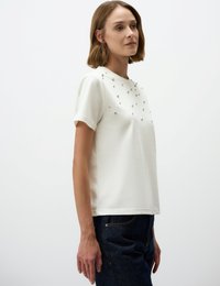 Kurzärmliges weißes T-Shirt mit Rundkragen und dekorativen silbernen Nieten, die in einem Muster auf der Vorderseite angeordnet sind. Lässige Passform mit glatter Textur.