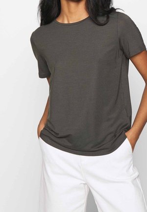 Person, die ein dunkelgraues kurzärmeliges T-Shirt und weiße Hose trägt, mit Händen in den Taschen, von Hals bis Hüfte gezeigt.