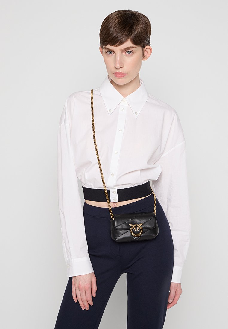 Sac en cuir noir à bandoulière avec quincaillerie dorée, design matelassé et sangle en chaîne. Le modèle porte une chemise blanche à boutons et un pantalon bleu marine.