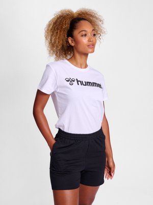 Kvinde med krøllet hår iført en hvid t-shirt med "hummel"-logo og sorte shorts, stående med hænderne i lommerne og kigger mod højre.