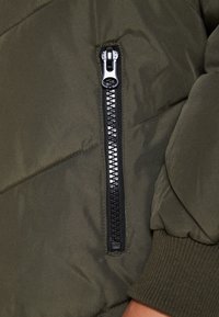 Detalhe de um casaco puffer verde-azeitona com um fecho éclair preto e ferragens prateadas, punhos canelados e padrão geométrico cosido no tecido.