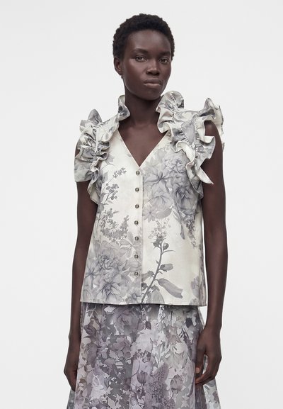 Γυναίκα που φοράει αμάνικο τοπ με κουμπιά και βολάν στους ώμους με λουλουδάτο σχέδιο, σε συνδυασμό με matching διαφανή φλοράλ φούστα.