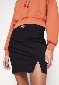Černá vroubkovaná mini sukně se bočním rozparkem a pasem s nášivkou "Tommy Jeans". Doplněna oranžovou mikinou.