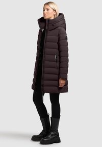 Bordeauxroter Steppmantel mit hohem Kragen, Reißverschluss vorne und Seitentaschen. Verfügt über ein gesteppte Muster und ist kombiniert mit schwarzen Leggings und Kampfstiefeln.