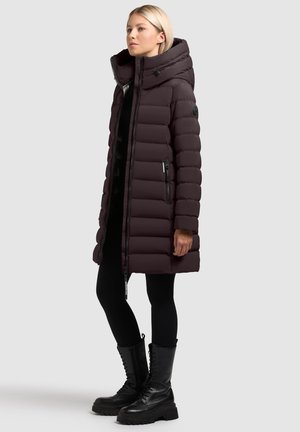 Abrigo acolchado marrón oscuro con cuello alto, cierre de cremallera frontal y bolsillos laterales. Presenta un diseño acolchado y se combina con leggings negros y botas de combate.