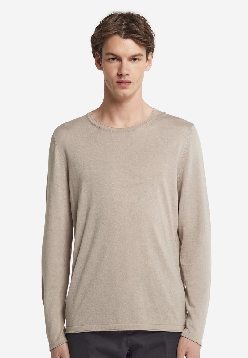 Giovane uomo che indossa una maglietta beige a manica lunga con collo tondo e pantaloni scuri, in piedi davanti a uno sfondo bianco semplice.