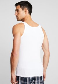 White cotton tank top with a ribbed texture, showing a fitted design and wide shoulder straps, worn by a model facing away from the camera.