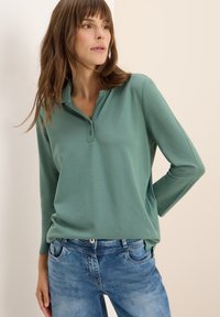 Polo verde con colletto a bottoni e maniche lunghe, abbinato a jeans sbiaditi blu chiaro. Tessuto liscio e vestibilità rilassata.