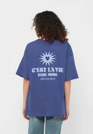 Oseba kodrastih las, oblečena v modro preveliko majico s srčnim in sončnim motivom ter napisom "C'EST LA VIE DARE MORE Life's too short."