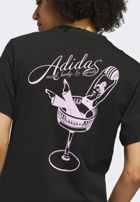 Čierne tričko s ružovou grafikou postavy relaxujúcej v pohári koktailu, so štylizovaným textom "Adidas mind, body, & spirits."