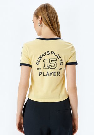 Camiseta amarilla con cuello y puños en azul marino, con un diseño de texto impreso en negro en la parte posterior: "SIEMPRE JUEGA A 15, GANAS JUGADOR."