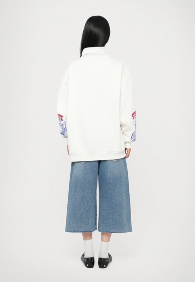 Fleece bianco oversize con colletto alto, dotato di grafiche rosse e blu sulle maniche. Abbinato a pantaloni shorts in denim a gamba larga di un blu medio.