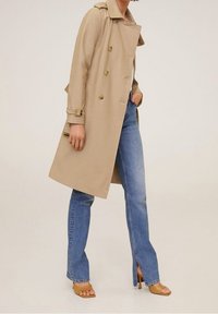 Kobieta w beżowym trenczu, jasnoniebieskich dżinsach z rozcięciem u dołu oraz beżowych sandałach na obcasie, stojąca na jednolitym tle.